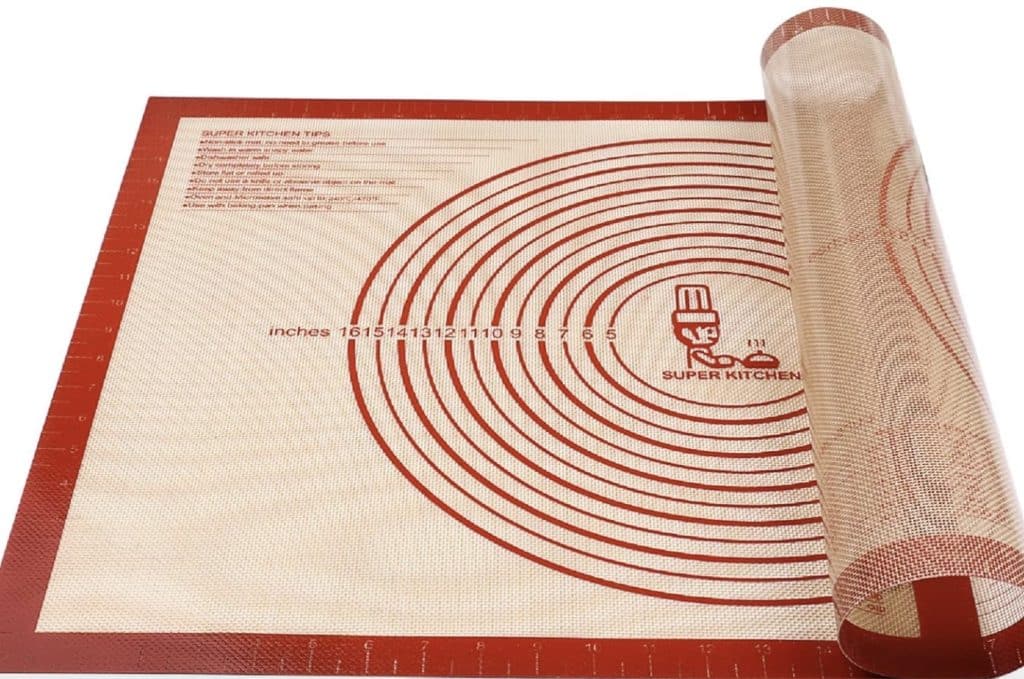 A red and beige silicone baking mat with measurement guides and concentric circles, perfect for whipping up the best Christmas cookies. Partially rolled at one corner, it features inch markings along the edges and a charming cartoon chef logo.